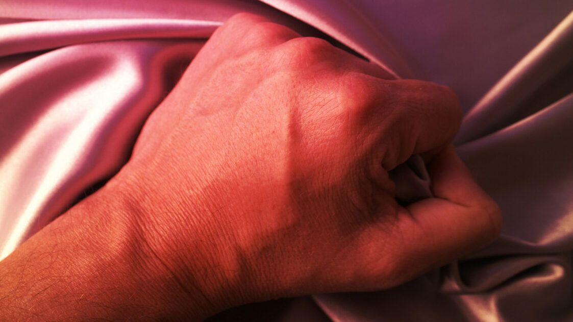 Mano cerrada sobre sábanas de satén rosa bajo luz cálida, imagen sugestiva para 5 tecnicas infalibles para volverlo adicto a ti en la cama.
