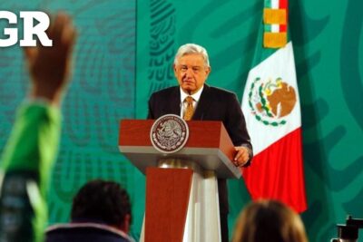 Hombre mayor de traje frente a un podio con el escudo y la bandera mexicana XY7aHl8x71E.