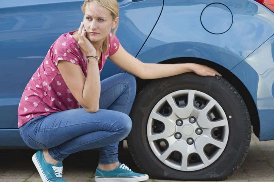 Mujer agachada junto a un coche con el neumático desinflado comprobando el daño y llamando por teléfono, aprende a reparar pinchazos en segundos con el kit antipinchazos para coche 1.