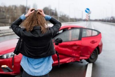 Mujer de espaldas con las manos en la cabeza frente a un auto rojo abollado en la carretera, imagen relacionada con cual es el significado de sonar con un accidente automovilistico 1
