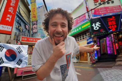 Hombre sonriente ante una calle con letreros en hangul y una bandera surcoreana que muestra la cultura coreana urbana.