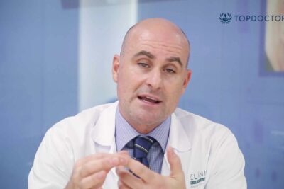 Médico con bata blanca y corbata realizando un gesto explicativo frente a fondo azul, db2aaaEeVM 2