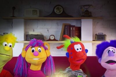 Cuatro marionetas coloridas sentadas en un sofá con fondo de estantería y ladrillo representando derechos humanos 1