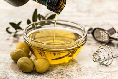 Cuenco de aceite de oliva vertiéndose junto a aceitunas verdes, descubre beneficios aceite oliva extra virgen.