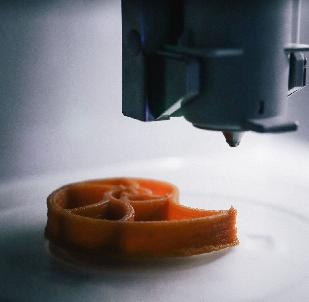 Descubre como funcionan las impresoras 3d de alimentos en segundos en un primer plano de la boquilla extruyendo una figura comestible naranja.