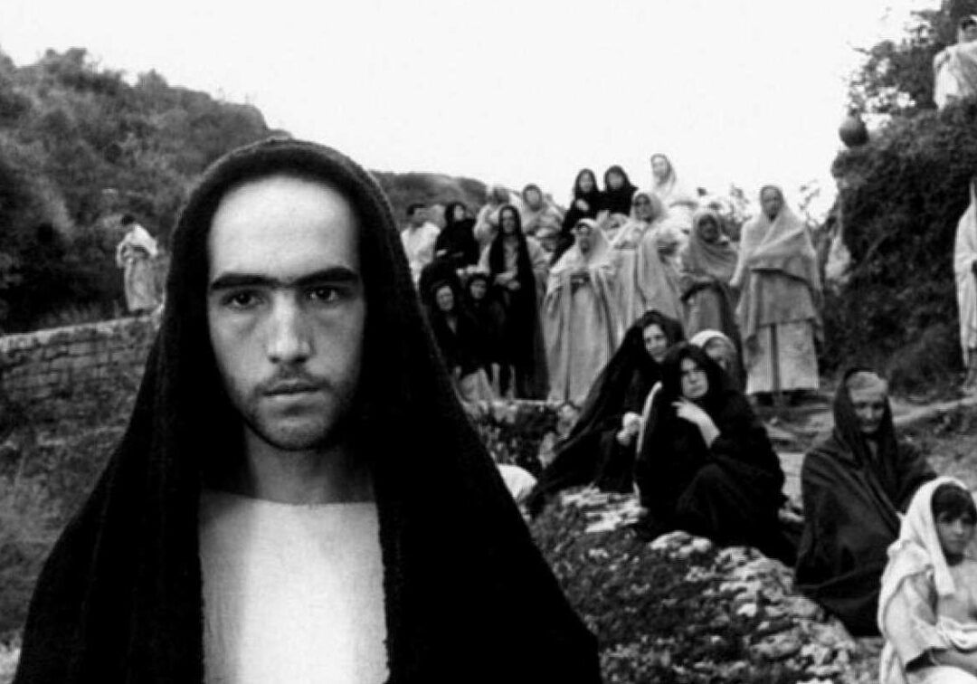Joven con capucha en primer plano frente a un grupo de personas con mantos y velos en actitud contemplativa, descubre el significado de la semana santa en tus propias palabras todo lo que necesitas saber