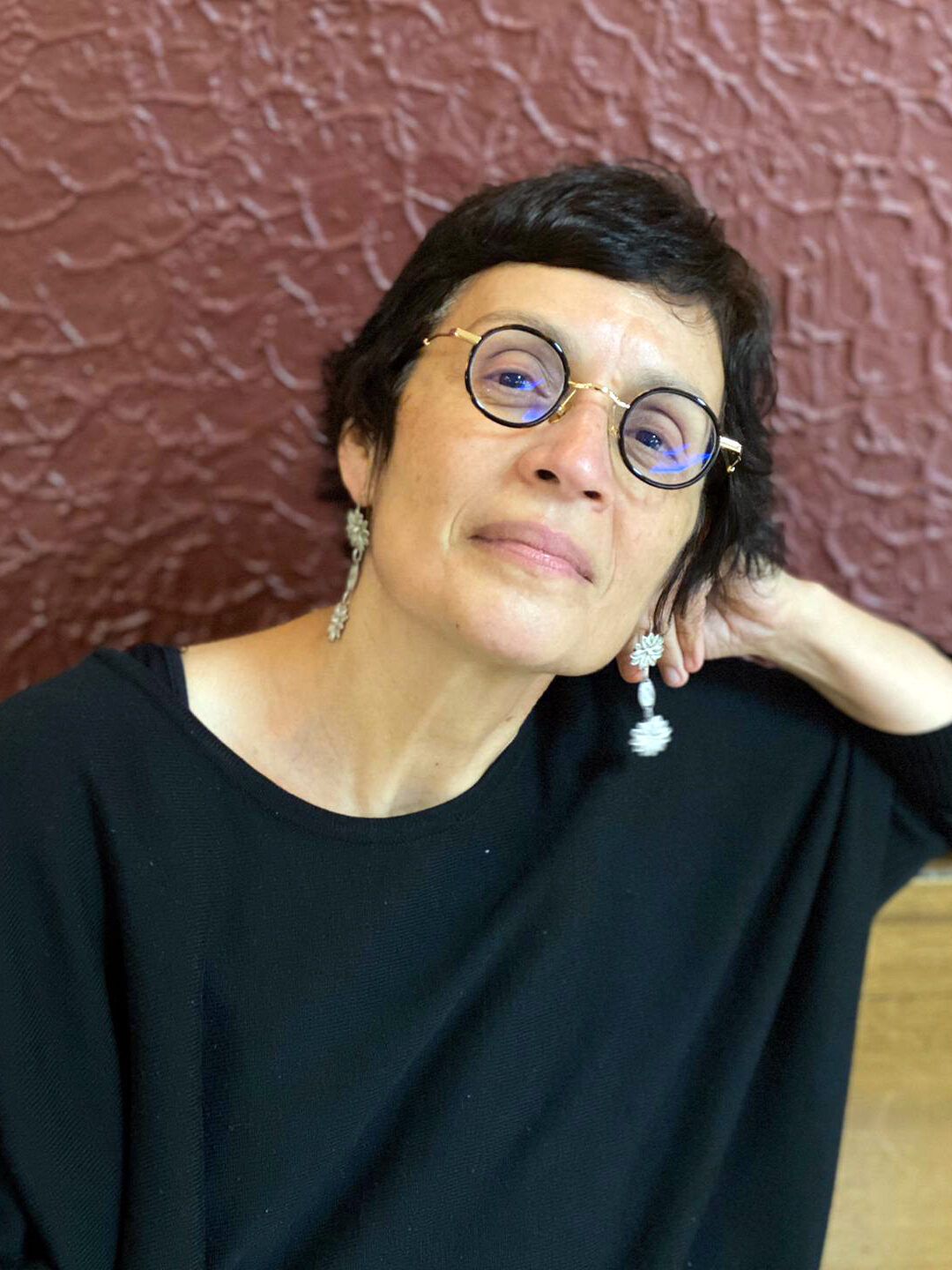 Mujer con gafas redondas y pendientes largos, mirada tranquila y postura relajada, descubre la belleza de la paciencia como quien espera el alba