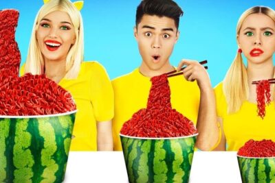 Tres personas con camisetas amarillas frente a mitades de sandía llenas de fideos rojos y palillos, descubre que comen los ninos cuando se deleitan con golosinas.