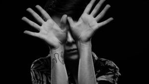 Retrato en blanco y negro de una persona cubriendo el rostro con las manos, primer plano del antebrazo con un tatuaje lineal visible; descubre significado tatuaje sol simbolismo