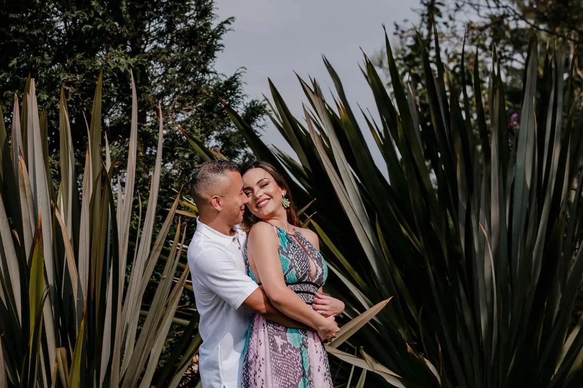 Pareja sonriente abrazada entre plantas en un jardín que evoca romance y celebración, descubre todo sobre el dia del amor y la amistad historia significado y tradiciones