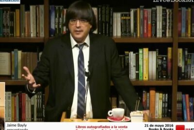 Persona dando una charla en una librería frente a estanterías repletas de libros y una mesa con micrófono y taza, descubre todo sobre que significa pecho frio