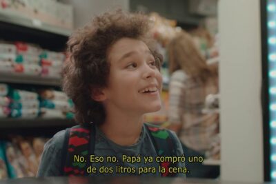 Niño sonriente en un pasillo de supermercado frente a estanterías de snacks y bebidas que evoca el impacto de las personas que comen dulces en la sociedad actual.