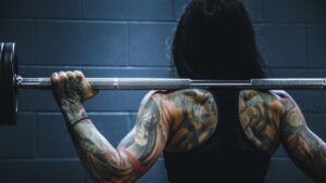 Persona de espaldas con tatuajes coloridos en brazos y espalda sosteniendo una barra de pesas, el significado de un tatuaje de angel descubre su simbolismo.