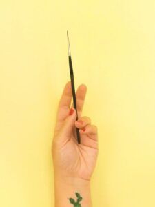 Mano con uñas rojas sosteniendo un pincel fino y tatuaje de planta en la muñeca, el simbolismo de un tatuaje de brujula y su significado 1