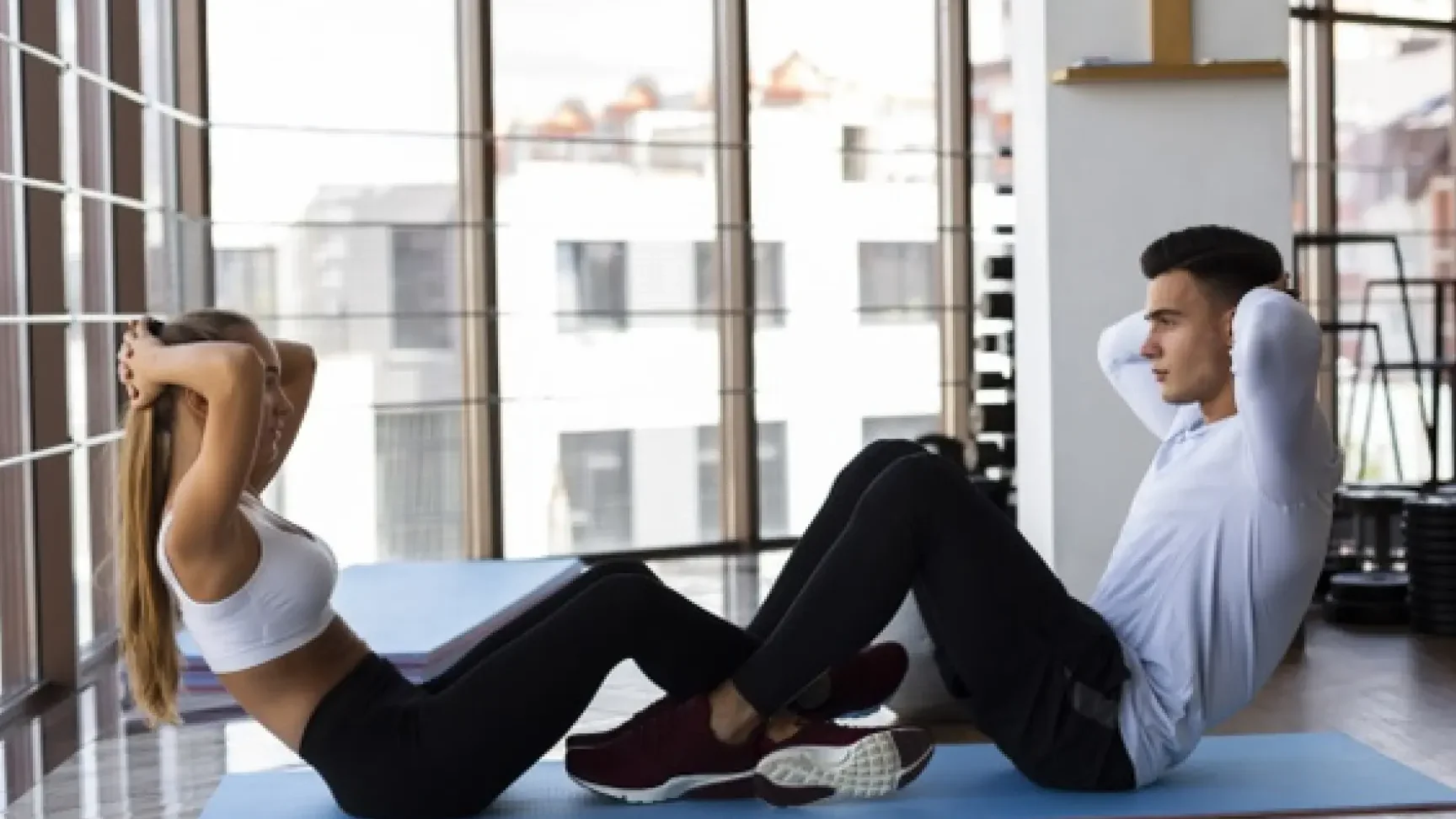 Dos personas realizan abdominales en colchonetas frente a grandes ventanales en un gimnasio, es saludable hacer ejercicio para abdomen todos los dias