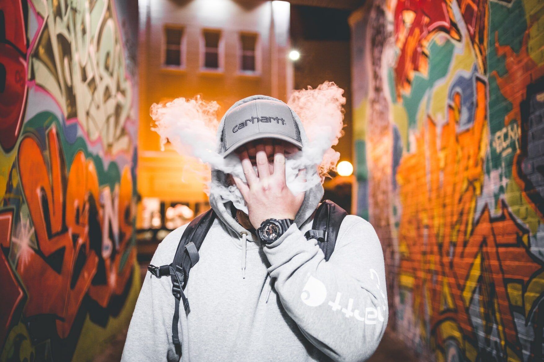 Persona con gorra y sudadera exhalando vapor frente a paredes con graffiti, es seguro fumar vapeo mientras se toman antidepresivos