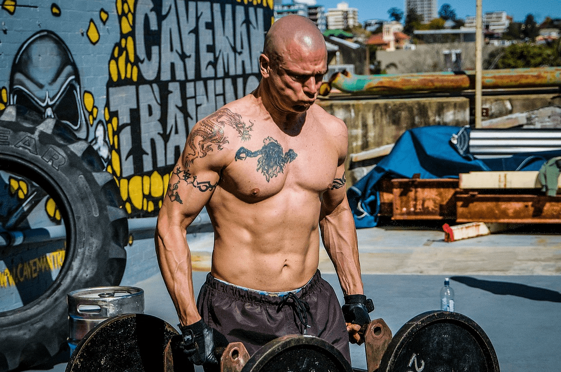 Hombre musculoso con tatuajes entrenando con pesas al aire libre, es seguro hacer abdominales dos veces al dia descubre la respuesta aqui