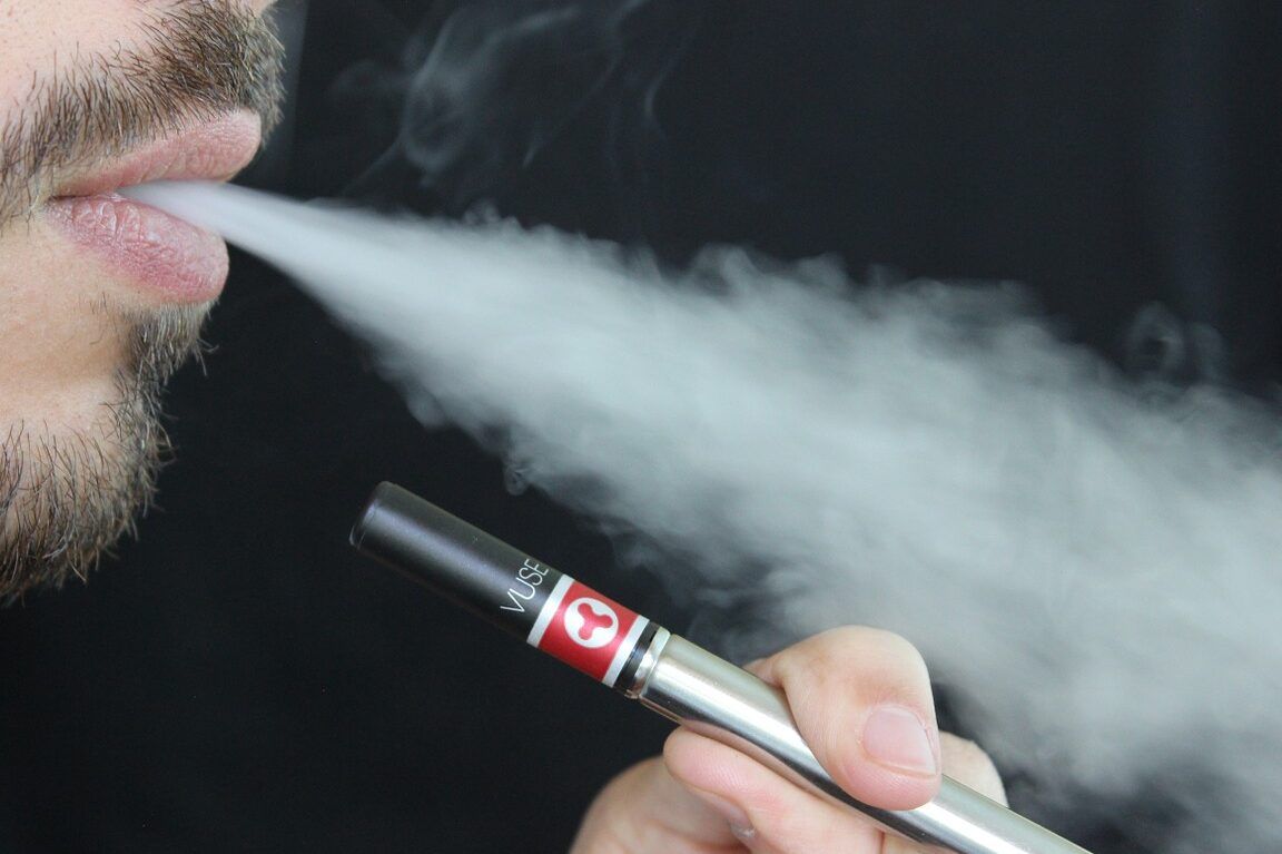 Primer plano de una persona exhalando vapor de cigarrillo electrónico; fumar con anginas oscuras descubre las consecuencias que podrias sufrir.