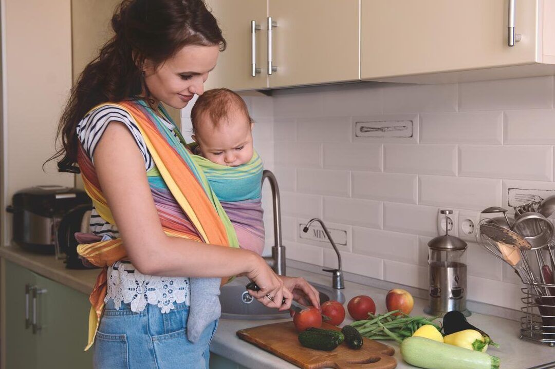Madre sostiene a su bebé en un fular mientras corta verduras en la cocina, fumar durante la lactancia descubre como afecta a tu bebe