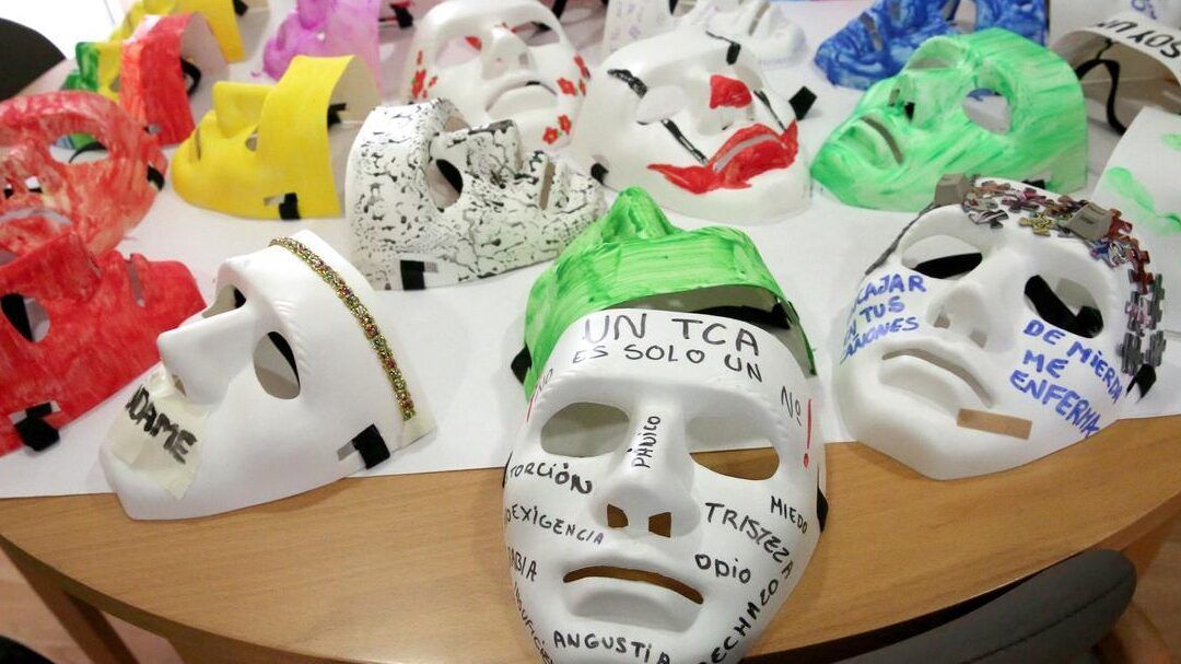 Máscaras decoradas con pintura y palabras sobre emociones y TCA en una mesa de taller, instituciones que luchan contra las falsas creencias cuales son y como lo hacen