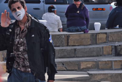 Hombre con rostro pintado saludando frente a un muro de piedra y personas sentadas, la importancia de no usar amoxicilina caducada 4 meses que riesgos implica