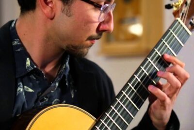 Músico tocando guitarra clásica con la mano en el mástil y camisa con estampado que evoca las abejas
