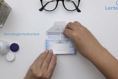 Manos abriendo un paquete de lentes contacto sobre una mesa con estuche, solución y gafas