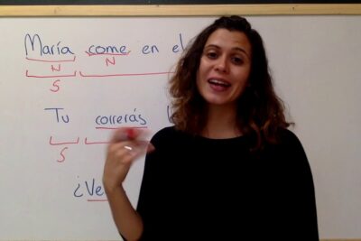 Instructora frente a una pizarra blanca con ejemplos de conjugación y anotaciones didácticas lhKYuxiWZKA