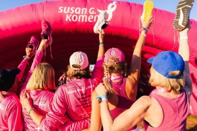 Participantes con camisetas y gorras rosas alzan zapatillas bajo un arco inflable en una caminata solidaria que logra awareness consideracion y conversion con tu publico objetivo.