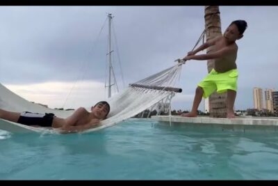 Dos niños jugando en una hamaca sobre la piscina junto a una palmera, escena de los willys