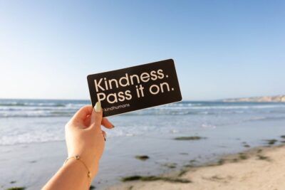Mano sosteniendo una tarjeta negra con el mensaje 'Kindness. Pass it on.' frente al mar promoviendo economia sostenible guia futuro prospero