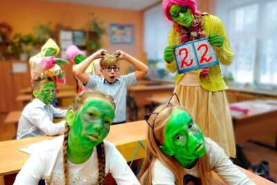 Niños y profesora disfrazados con caras pintadas de verde en un aula mientras una docente sostiene un cartel, que comen los patitos bebes descubre la alimentacion ideal para cuidar a estas adorables criaturas
