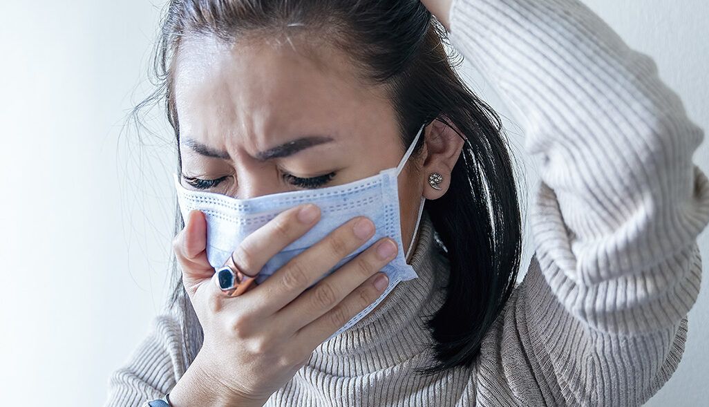 Mujer tosiendo con mascarilla: que consecuencias tiene dar positivo en covid despues de 10 dias y qué observar, primer plano de mujer cubriéndose la boca con la mano y mostrando malestar por la tos.