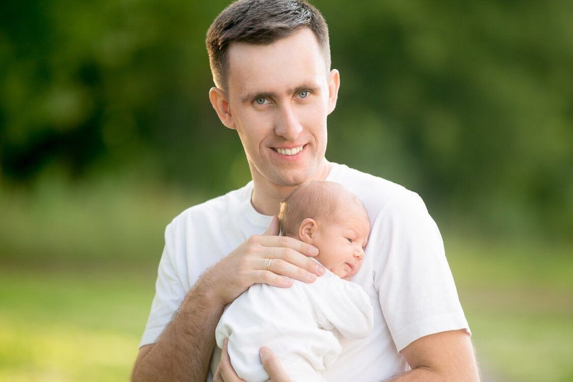 Padre sonriente sostiene a un recién nacido en brazos en un entorno natural, que significa ser un bebe no binario descubre todo aqui