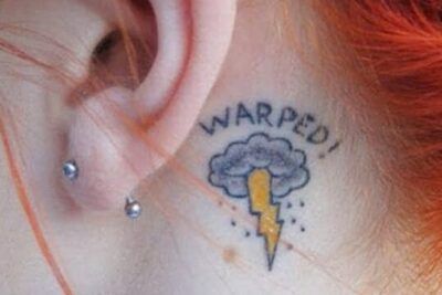 Tatuaje pequeño detrás de la oreja de una nube con rayo amarillo y texto "WARPED!" que significa un tatuaje de nube guia completa y significativa 1