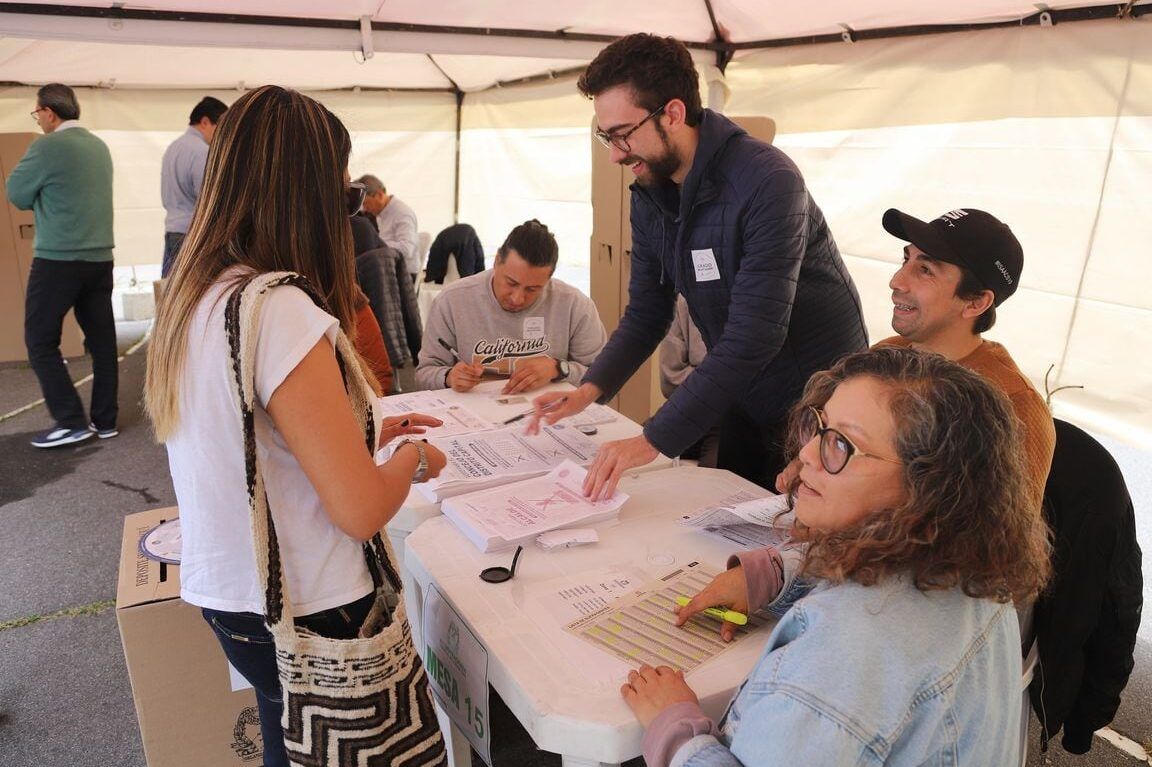 Personas interactuando en una mesa electoral dentro de una carpa donde se registran votantes y se entregan papeletas; que sucede si en un pais gana el voto en blanco descubre las consecuencias aqui