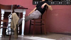 Persona reclinada en una silla con pantalones estampado de leopardo junto a una mesa y un bolso, significado crona historia simbolismo.