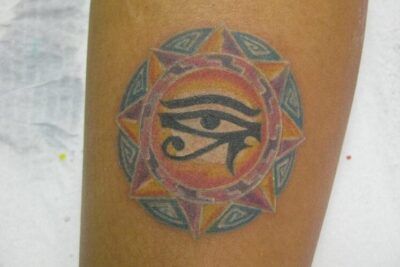 Tatuaje en antebrazo con un ojo central dentro de un sol geométrico de tonos naranjas, morados y azules, significado de un tatuaje de luna descubre su simbolismo.
