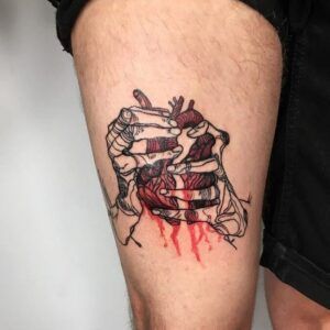 Tatuaje en muslo de manos esqueléticas que abren costillas mostrando un corazón anatómico sangrante significado del tatuaje de corazon roto descubre mas aqui