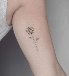 Tatuaje minimalista en tinta negra de una rosa con tallo caligráfico que ilustra el significado y simbolismo de un tatuaje de rosa blanca.