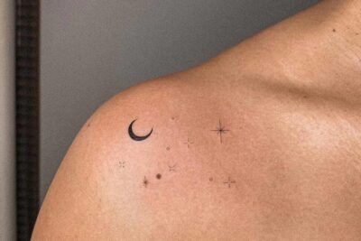 Tatuaje minimalista de media luna y pequeñas estrellas sobre la clavícula que evoca el simbolismo de un tatuaje de luna y estrella en el tobillo